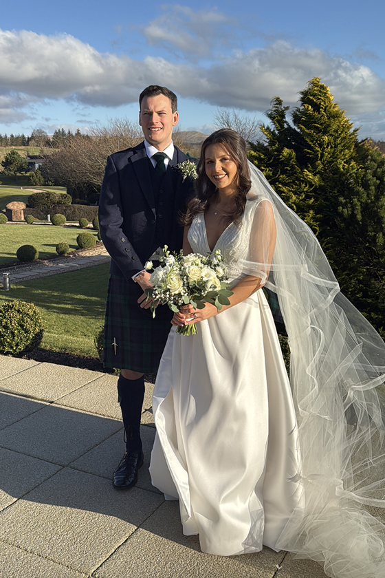 Bride holding bouquet stands smiling with her groom outside wedding venue